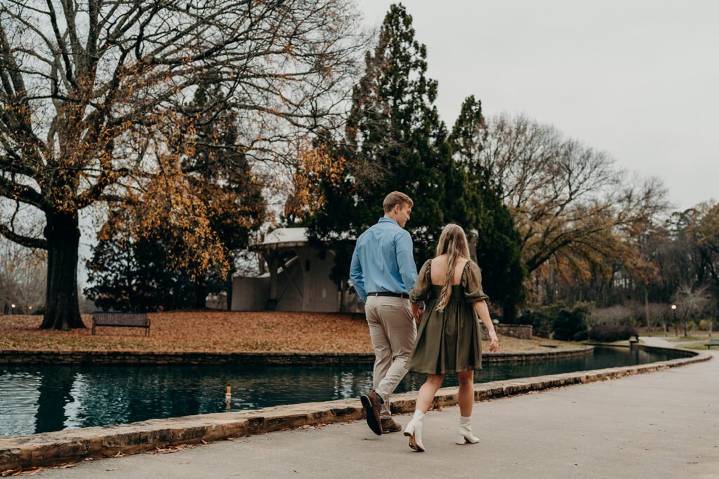 Photo of couple walking holding hands through park next to a blue pond while the trees change colors for fall