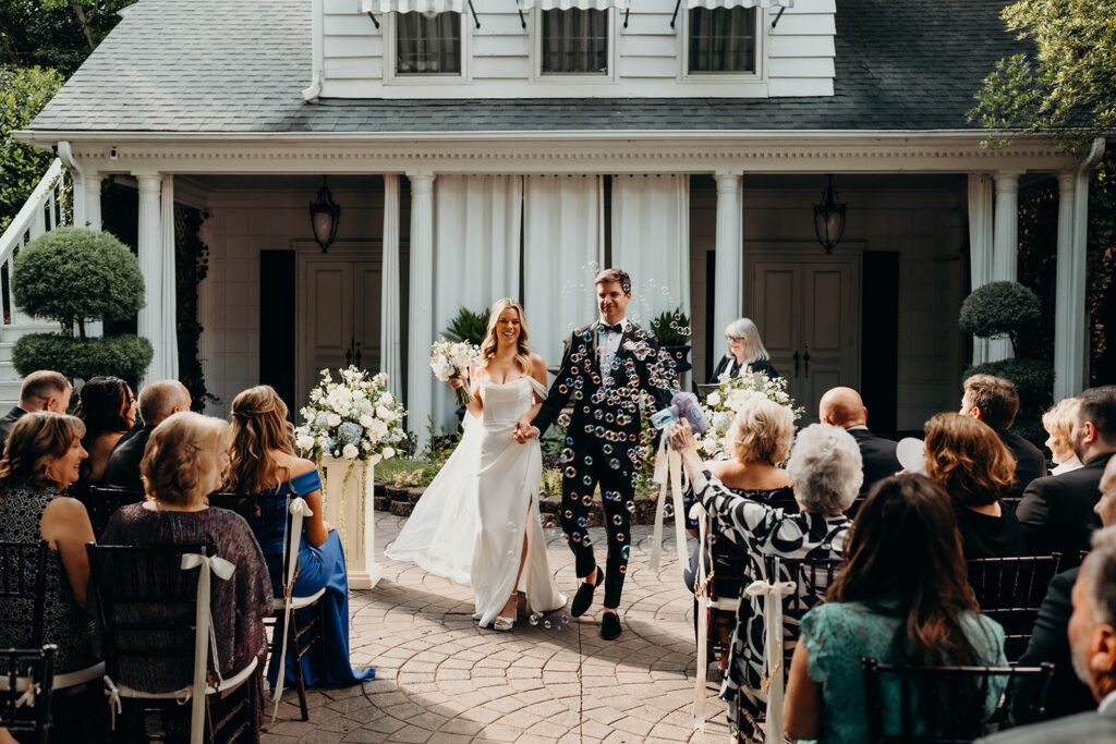 Summer wedding ceremony in secret private courtyard on a sunny day with lush greenery and blue flowers walking down aisle