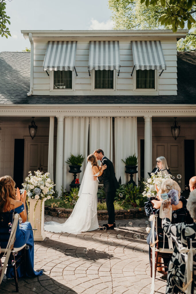 Summer wedding ceremony in secret private courtyard on a sunny day with lush greenery and blue flowers walking down aisle