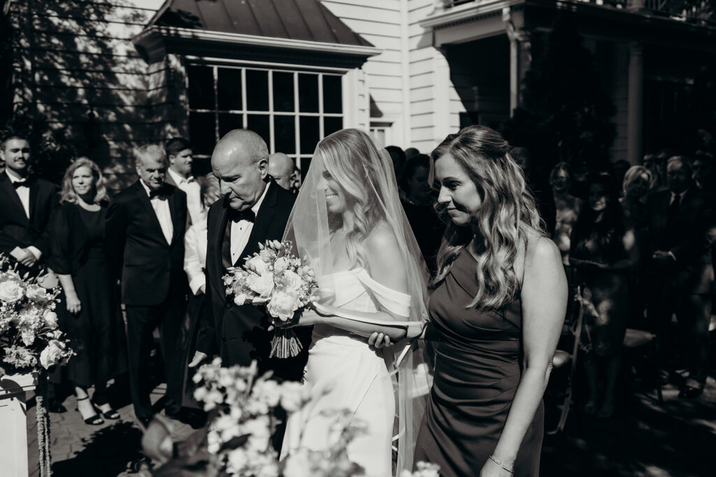 Summer wedding ceremony in secret private courtyard on a sunny day with lush greenery and blue flowers walking down aisle