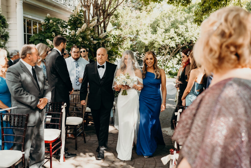 Summer wedding ceremony in secret private courtyard on a sunny day with lush greenery and blue flowers walking down aisle
