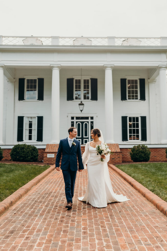 bride and groom walking in front of the timeless white estate with big columns and white exterior at their rose hill estate wedding