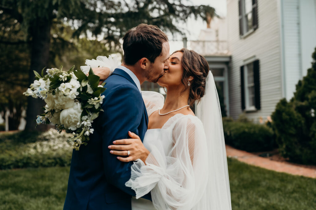 bride and groom kissing in front of the estate while she drapes her flowers over his neck at their rose hill estate wedding