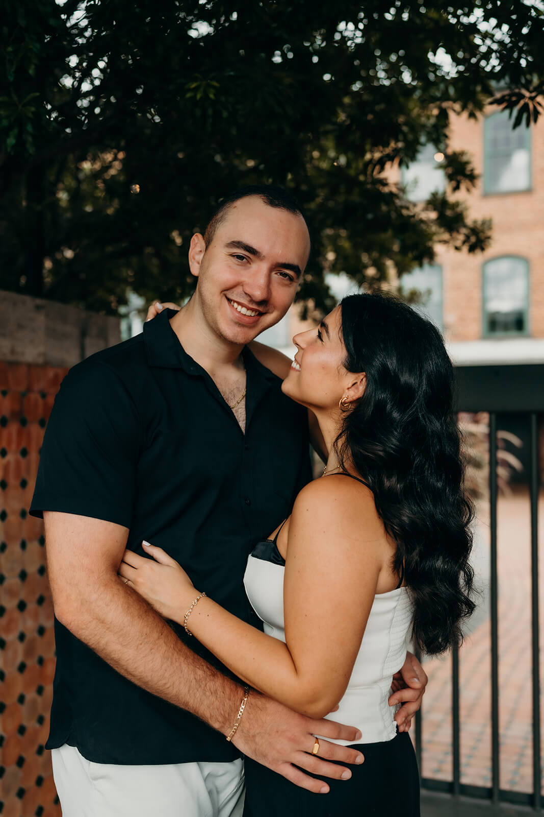 Close up photo of the couple where they are holding each other while he looks at the camera and she is smiling at him